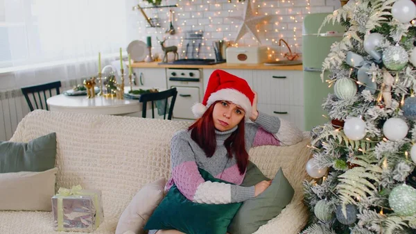 Kvinna i tomte hatt sitter på soffan nära julgran och kliar huvudet. Missnöjd och omtänksam kvinna tittar på kameran, reflektera över kommande helgdagar — Stockfoto
