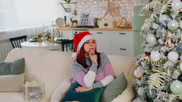 Jeune femme au chapeau de Père Noël assise sur le canapé près de l'arbre de Noël dans le salon. Robinets féminins réfléchis menton avec le doigt, penser aux vacances à venir. — Photo