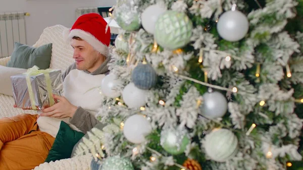 Homem em Papai Noel com caixa de presente sentado no sofá perto da árvore de Natal. O macho satisfeito considera e abraça Natal presente no apartamento com árvore decorada festiva. Conceito de surpresa durante as férias. — Fotografia de Stock