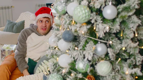 Triste homme au chapeau de Père Noël assis sur le canapé près de l'arbre de Noël dans le salon. Malheureux mâle regarde arbre décoré, enlève son chapeau de Noël et le jette. — Photo
