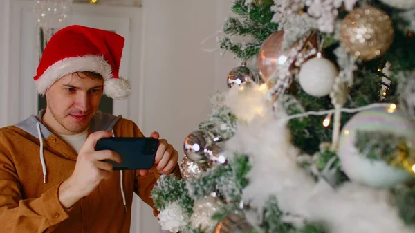 Mann fotografiert Weihnachtsbaum. Junger Mann mit Weihnachtsmütze fotografiert auf Handy Weihnachtsbaum mit Lichterketten und Christbaumkugeln in den Neujahrsferien — Stockfoto