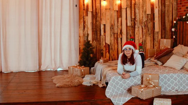Женщина лежит на кровати с подарками. Полное тело положительной женщины в шляпе Санты и теплые носки, лежащие на кровати среди обернутых рождественских подарков в уютной студии под светящимися лампами и глядя в камеру — стоковое фото
