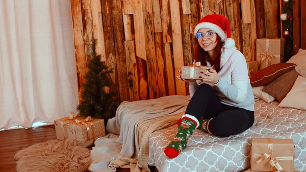Улыбающаяся женщина с рождественским подарком. Молодая улыбающаяся женщина в тёплой одежде Санты и очках сидит на кровати с обернутой рождественской подарочной коробкой в студии — стоковое фото
