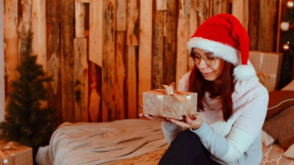 Улыбающаяся брюнетка в новогодней шляпе с завернутыми подарками. Счастливая женщина держит подарок и пытается угадать, что внутри. — стоковое фото