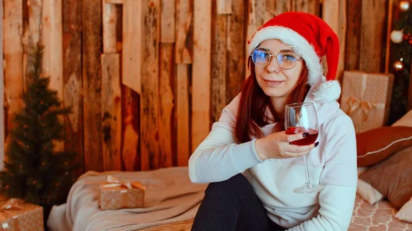 在圣诞礼物附近喝酒的女人。在庆祝新年的时候，戴着眼镜、头戴圣诞礼帽的女性正坐在床上，喝着一大杯红酒。坐在包装礼品盒旁的女人 — 图库照片