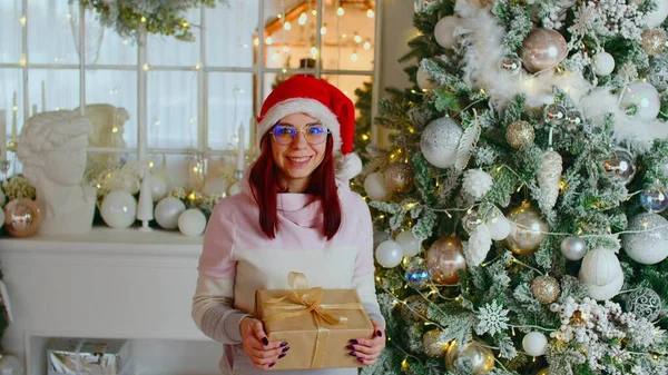 Lycklig kvinna med julklappslåda. Positiv ung kvinna i Santa hatt och glasögon stående med presentförpackning bunden med band nära julgran dekorerad med kransar och grannlåt — Stockfoto