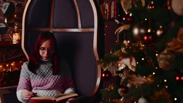 Koncentrerad kvinna i varm tröja och glasögon läsa bok på fritid nära julgran dekorerad med gloving girlanger i mysig lägenhet — Stockfoto
