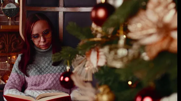 Сконцентрированная женщина в теплых свитерах и очках читает книгу в свободное время возле елки, украшенной перчаточными гирляндами в уютной квартире — стоковое фото