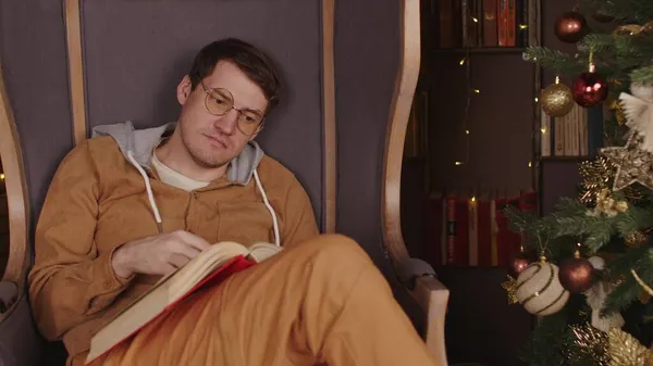 Hombre leyendo un libro cerca del árbol de Navidad. Hombre concentrado en jersey cálido y anteojos lectura libro en tiempo libre cerca del árbol de Navidad en acogedor apartamento. — Foto de Stock