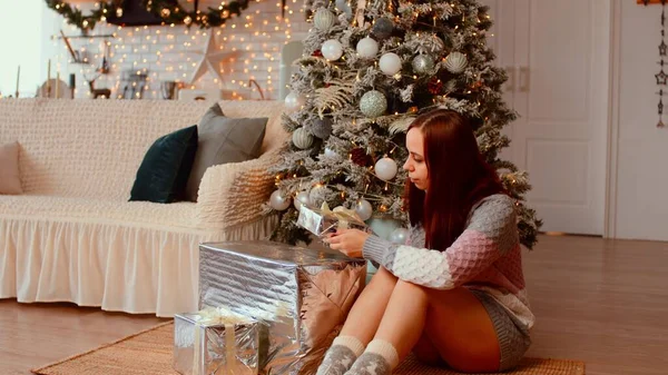 Женщина под елкой с подарками. Женский свитер сидит на полу под праздничной елкой рядом с диваном и берет обернутую подарочную коробку из кучи в уютной квартире, украшенной гирляндами — стоковое фото