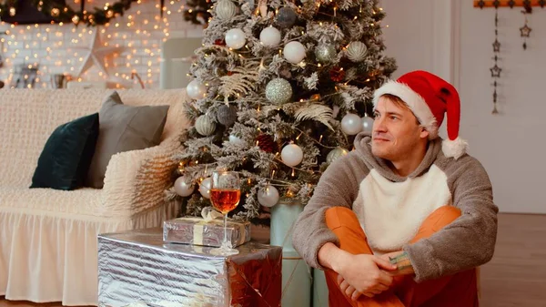 Gelukkige man in kerstmuts zit op de vloer in de buurt van de kerstboom in de woonkamer. jong mannetje wegkijkend dromerig en glimlachend. — Stockfoto