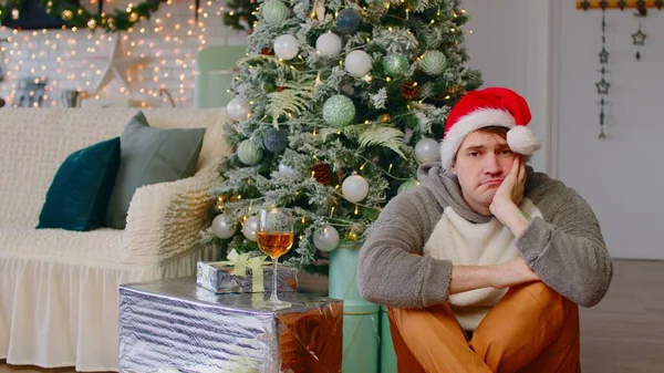 Homem triste em Papai Noel sentado no chão perto da árvore de Natal na sala de estar. Homem solitário suspira, olhando para a câmera, em seguida, para baixo durante as férias comemorativas. — Fotografia de Stock