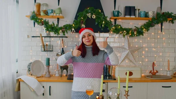 Молодая женщина в шляпе Санты и свитере, показывающая большие пальцы вверх, стоящая на праздничной кухне. Красивая женщина смотрит в камеру, улыбается и показывает жест одобрения. — стоковое фото