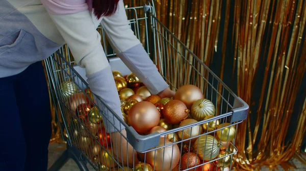 Donna con carretto pieno di bagattelle d'oro per Natale. Vista laterale del raccolto anonimo femminile scegliendo palline decorative d'oro nel carrello per la preparazione del Natale mentre — Foto Stock