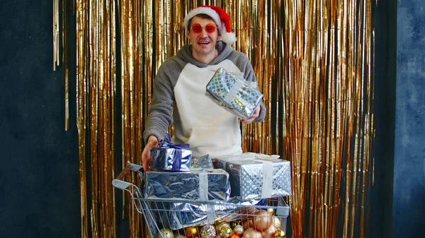 Jeune homme dans des lunettes lumineuses et chapeau de Père Noël debout avec chariot rempli de boules assorties et enveloppé cadeaux de Noël contre le mur avec tinsel chatoyant. — Photo