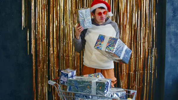 Jeune homme dans des lunettes lumineuses et chapeau de Père Noël debout avec chariot rempli de boules assorties et enveloppé cadeaux de Noël contre le mur avec tinsel chatoyant. — Photo