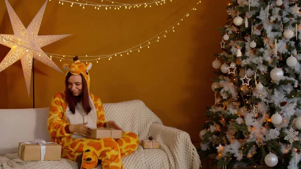 Kvinna i pyjamas med presentaskar sittande på soffan nära julgranen. Ung kvinna överväger och skakar julklappar i förväntan. Begreppet överraskningar under semestern. — Stockfoto
