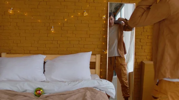 Vista trasera de la cosecha masculina mirando en el espejo y poniéndose camisa amarilla cerca de la cama con bola disco en la manta en el dormitorio decorado con luces de hadas para Navidad —  Fotos de Stock
