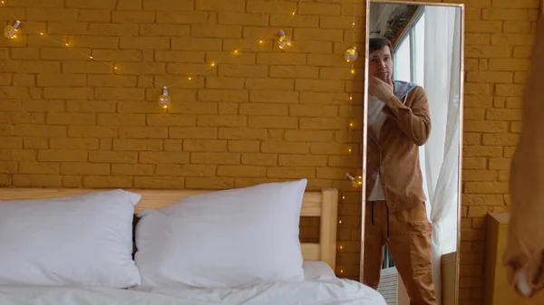 Mężczyzna ubiera się w sypialni ozdobiony świecącą girlandą. Widok z tyłu upraw samiec patrząc w lustro i wprowadzenie żółtej koszuli w pobliżu łóżka z kulą disco na kocu — Zdjęcie stockowe