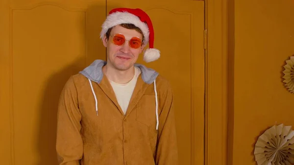 Hombre feliz en sombrero de santa y gafas de moda expresa emociones positivas y festivas sobre fondo marrón. Elegante hombre mirando a la cámara y sonriendo, de pie en la habitación. — Foto de Stock