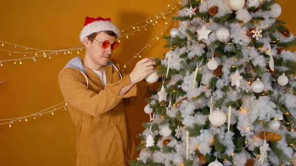 Man in santa hat and fashionable glasses decorating Christmas tree at home. Young male hanging white bauble on fir branch while preparing for Christmas celebration at home. — Stock Photo, Image