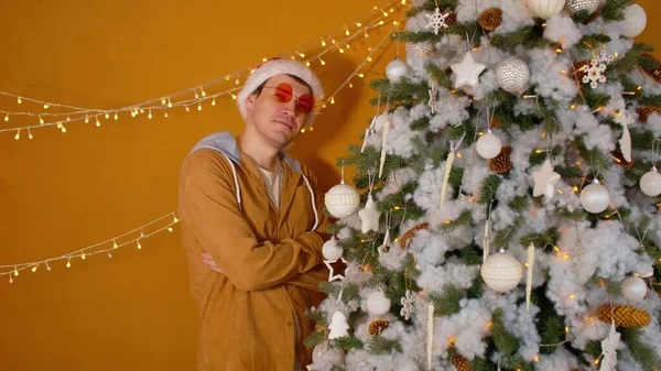 Junger Mann mit Weihnachtsmannmütze steht mit verschränkten Armen am Weihnachtsbaum und blickt in die Kamera. Schöner Mann mit modischer Brille lächelt und posiert neben geschmücktem Baum. — Stockfoto