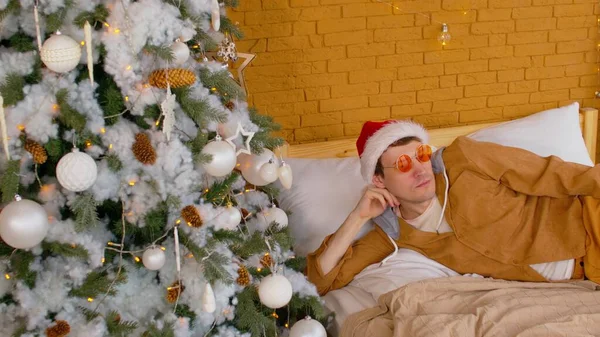 Молодий чоловік у капелюсі Санта лежить на ліжку біля ялинки і озирається навколо. Розслаблений самець в модних окулярах відпочиває і насолоджується затишною святковою атмосферою . — стокове фото
