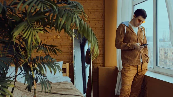 Koncentrerad ung man i ljusa snygga glasögon står nära fönstret och sms: a på mobiltelefon i sovrum med handflata och dekorativa glödande krans för julhelgen — Stockfoto