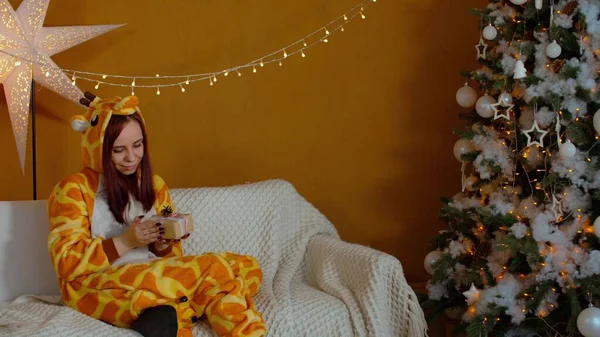 Femme en pyjama avec boîte cadeau assise sur le canapé près de l'arbre de Noël. Jeune femme considère et secoue Noël présent en anticipation. Concept de surprise pendant les vacances. — Photo