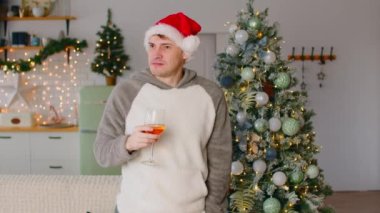 Noel Baba şapkalı bir adam bir bardak içecekle oturma odasında Noel ağacının yanında duruyor. Düşünceli erkek etrafına bakar, çenesini kaşır, parmaklarını sayar, yaklaşan tatilleri düşünür..