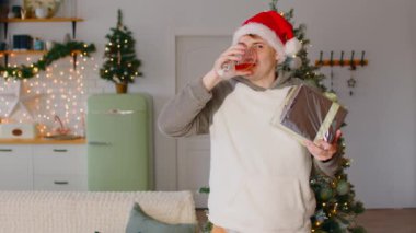 Noel arifesinde, elinde bir hediye ve bir bardak şarap olan bir adam. Noel Baba şapkalı mutlu bir adam elinde bir Noel hediyesi tutuyor ve bir Noel ağacının arka planında şarap içiyor.