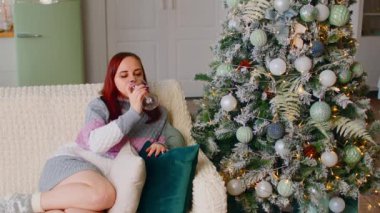 Noel ağacının yanındaki kanepede oturan ve alkollü içecek içen bir kadın. Alkolle dinlenen uzun süveterli tatmin olmuş bir kadın ve dairede Noel 'i süslü bir ağaçla kutluyor..