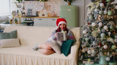 Noel Baba şapkalı genç bir kadın oturma odasındaki Noel ağacının yanındaki koltukta oturuyor. Mutlu kadın Noel hediyesini düşünür ve kucaklar. Tatillerde sürpriz kavramı.