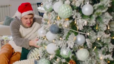 Genç adam kanepede oturuyor ve Noel ağacına bakıyor. Hayalperest erkek, parlak çelenklerle süslenmiş stüdyo dairesinde oturup Noel ağacı süslemelerini düşünür ve ona dokunur..