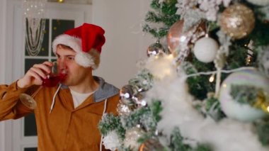 Noel ağacının yanında şarap içen adam. Kırmızı Noel Baba şapkalı bir erkek camdan kırmızı şarap içerken Noel ağacının yanında yeni yılı kutluyor.