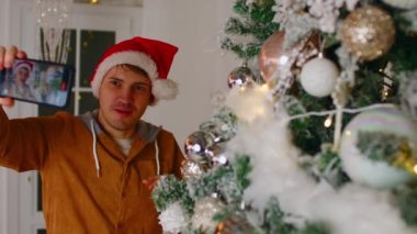 Noel Baba şapkalı, cep telefonlu genç adam Noel ağacının yanında duruyor. Pozitif erkek cep telefonu için tebrik videosu çekiyor. Tatilleri evde kutlama kavramı.