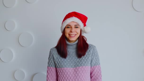 Jonge vrouw in santa hoed op de achtergrond van witte patroon muur. Portret van mooie vrouw in trui kijkend naar camera en glimlachend. — Stockvideo