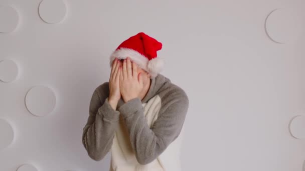 Man in de kerstman hoed verbergt gezicht met handen op de achtergrond van witte patroon muur. Grappig mannetje flirten en kijken uit handen op camera. — Stockvideo