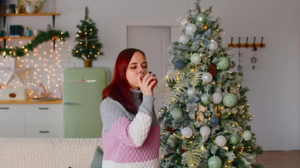 Jovem mulher bebendo bebida e dançando perto da árvore de Natal na sala de estar. Entretenimento feminino com copo de álcool durante as férias de celebração. — Vídeo de Stock
