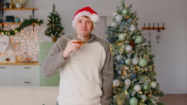 Ο άνθρωπος με το καπέλο Σάντα πίνει κρασί κοντά στο χριστουγεννιάτικο δέντρο στο σαλόνι. Νεαρό αρσενικό πίνει λευκό κρασί από γυαλί, ενώ γιορτάζει την Πρωτοχρονιά κοντά διακοσμημένο δέντρο στο σπίτι. — Αρχείο Βίντεο