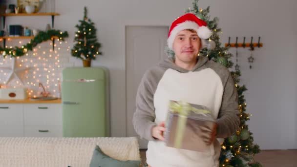 Happy man in santa hat dancing with gift box in hands near Christmas tree in living room. Positive male looking at camera, smiling and dancing while celebrating holidays near decorated tree at home. — Stock Video