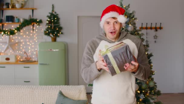 Ο άνθρωπος λαμβάνει χριστουγεννιάτικο δώρο από την κάμερα. Νεαρό αρσενικό σε Σάντα καπέλο στέκεται στο διαμέρισμα διακοσμημένα με εορταστική χριστουγεννιάτικο δέντρο και χαρά τυλιγμένο κουτί δώρου δεμένα με κορδέλα. — Αρχείο Βίντεο