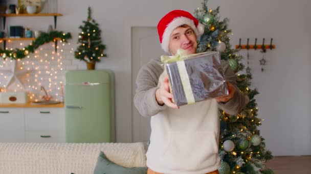 Man geeft kerstcadeau aan de camera. Jonge man in Santa hoed staan in appartement versierd met feestelijke kerstboom en reiken uit verpakt geschenkdoos gebonden met lint — Stockvideo