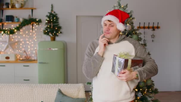 Человек в шляпе Санты с подарочной коробкой, стоящей возле елки в гостиной. Задумчивый мужчина смотрит вокруг, царапает подбородок, размышляет о подарках и предстоящих праздниках. — стоковое видео