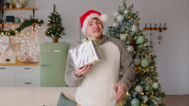 Молодой человек держит завернутый подарок. Улыбающийся человек в новогодней шляпе, держащий завернутый подарок и смотрящий в камеру на фоне новогодней елки — стоковое видео