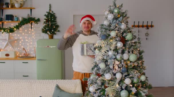 Mano masculina saludando cerca del árbol de Navidad. Feliz joven con ropa de abrigo y sombrero de Santa sonriendo y saludando de la mano mientras está de pie cerca del árbol de Navidad con caja de regalo en casa — Vídeos de Stock