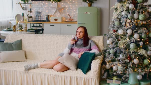 Frau sitzt auf Sofa neben Weihnachtsbaum und trinkt alkoholisches Getränk. Zufriedene Frau im langen Pullover, mit Alkohol in der Hand und Silvester in Wohnung mit festlich geschmücktem Baum. — Stockvideo