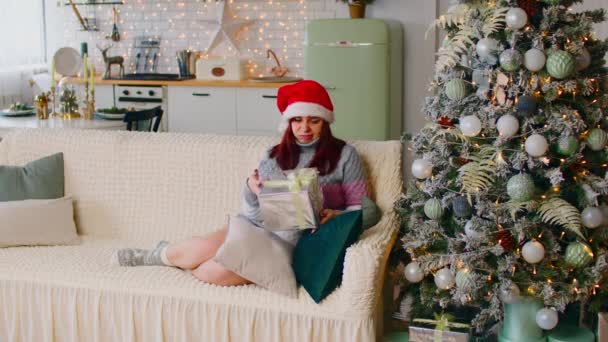 サンタの帽子でいらいらした女性の完全な体と暖かい服座ってソファの上に悪いラッププレゼント近くのクリスマスツリーでスタジオアパートで輝くガーランドで飾られています — ストック動画