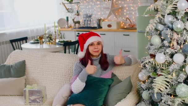 一个大拇指的女人坐在装饰过的圣诞树旁的沙发上，一头长长的红头发、头戴温暖毛衣、头戴圣诞帽的积极的年轻女性，竖起大拇指，看着相机 — 图库视频影像