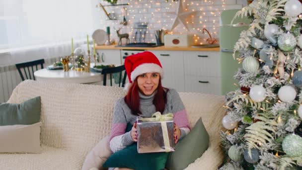 Woman in santa hat with gift box sitting on sofa near Christmas tree. Satisfied female considers christmas present in apartment with festive decorated tree. Concept of surprise during holidays. — Stock Video